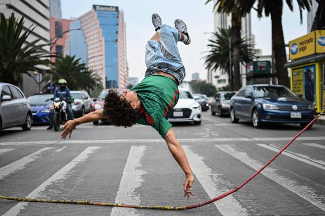 Venezuelan immigrant Edwin Bravo, wearing a Mexico national football team jersey, performs an acrobatic move while jumping a rope held by two companions as they collect money from drivers waiting at a traffic light in Mexico City on June 19, 2026. (Photo by Yuri CORTEZ / AFP)