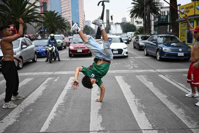 Venezuelan immigrant Edwin Bravo (C), wearing a Mexico national football team jersey, performs an acrobatic move while jumping a rope held by two companions as they collect money from drivers waiting at a traffic light in Mexico City on June 19, 2026. (Photo by Yuri CORTEZ / AFP)