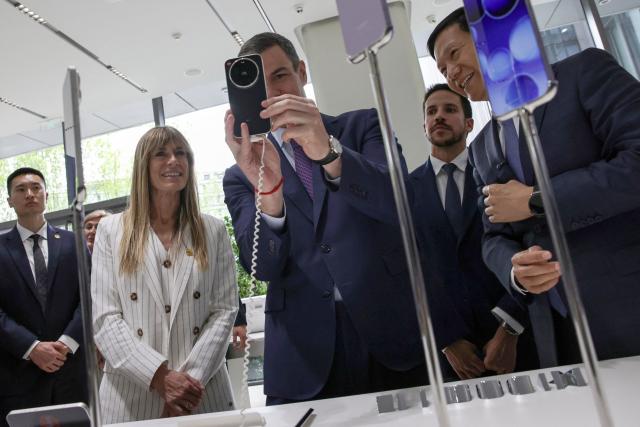 (FILES) Spain's Prime Minister Pedro Sanchez (C) takes a photo with a Xiaomi smartphone in presence of his wife Begona Gomez (2L) and Founder, Chairman & CEO of Xiaomi, Lei Jun (R), at the Xiaomi headquarters in Beijing on April 13, 2026. A Spanish court has ordered the wife of Prime Minister Pedro Sanchez, Begona Gomez, to stand trial for corruption and banned her from leaving the country, according to a court order released on June 20, 2026. Judge Juan Carlos Peinado ordered Gomez to hand in her passport and to check in with the authorities twice a month until a verdict is reached in the case, the ruling said. (Photo by ANDRES MARTINEZ CASARES / POOL / AFP)