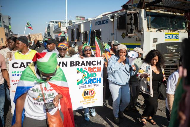 A group of about 100 people, some carrying banners and waving South African flags, take part in a march, organised by the March and March movement, protesting against illegal immigrants, in Wynberg, a Cape Town suburb, on June 20, 2026. (Photo by RODGER BOSCH / AFP)