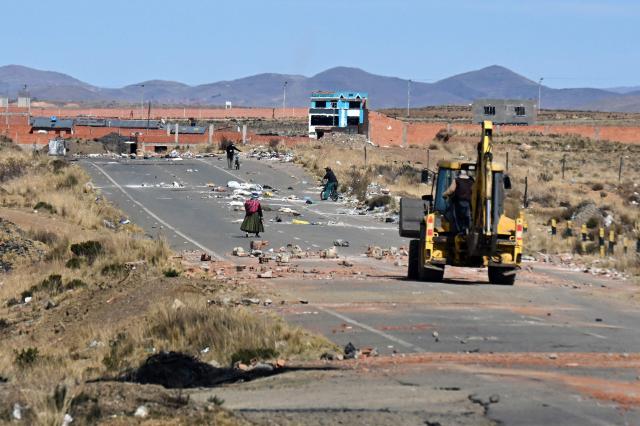 A woman walks in the highway as a backhoe bucket clears the roadblock in Mazo Cruz, La Paz department, Bolivia on June 20, 2026, after Bolivian President Rodrigo Paz declared state of emergency. A woman walks amid rubble as a a Bolivia's president declared a state of emergency and deployed police and bulldozers to clear anti-government roadblocks that have paralyzed the Andean nation for more than six weeks. An array of anti-government unions, Indigenous groups and coca farmers have marched through cities and blocked main roads across the country with rubble, logs and debris. (Photo by AIZAR RALDES / AFP)
