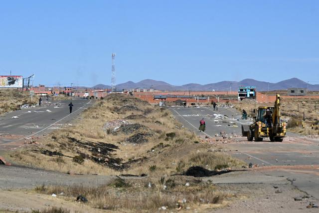 A woman walks in the highway as a backhoe bucket clears the roadblock in Mazo Cruz, La Paz department, Bolivia on June 20, 2026, after Bolivian President Rodrigo Paz declared state of emergency. Bolivia's president declared a state of emergency and deployed police and bulldozers to clear anti-government roadblocks that have paralyzed the Andean nation for more than six weeks. An array of anti-government unions, Indigenous groups and coca farmers have marched through cities and blocked main roads across the country with rubble, logs and debris. (Photo by AIZAR RALDES / AFP)
