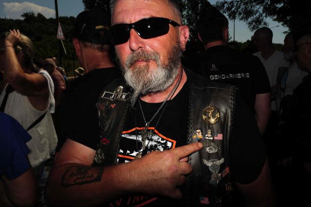 An attendee gestures to his vest pointing to a pin referencing Coluche as he takes part in a "Run Coluche" biker gathering as part of a tribute to French comedian and actor Coluche 40 years after his death, in Opio, southeastern France, on June 21, 2026. (Photo by Valery HACHE / AFP)