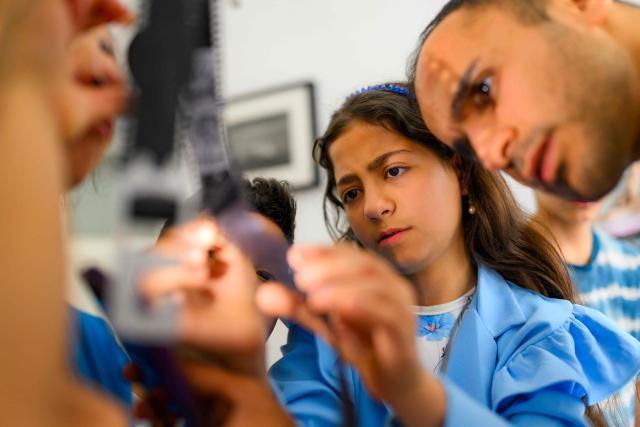 Syrian photographer, educator and co-founder of the project Serbest Salih (R) works with children during a workshop held as part of Fotohane Darkroom project in Mardin, southeastern Turkey, on June 13, 2026. In a dimly lit room illuminated by a pair of red lamps, eight-year-old Zeynep waits to see the photographs she has taken, now trapped as shadows and silhouettes on a roll of film. Originally from the southeastern province of Mardin, Zeynep is among eight children taking part in a two-month analog photography workshop for local and migrant youth near Turkey's borders with Iraq and Syria. The project, called Fotohane Darkroom, started in Mardin in 2024, by Kilic and Syrian photographer and educator Serbest Salih. In Turkish, Arabic, Kurdish and Persian, Fotohane means "house of photo", a name chosen by the children. (Photo by Yasin AKGUL / AFP)