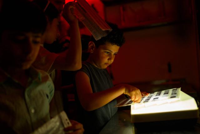 Children look at camera films in a darkroom during a workshop held as part of Fotohane Darkroom project in Mardin, southeastern Turkey, on June 14, 2026. In a dimly lit room illuminated by a pair of red lamps, eight-year-old Zeynep waits to see the photographs she has taken, now trapped as shadows and silhouettes on a roll of film. Originally from the southeastern province of Mardin, Zeynep is among eight children taking part in a two-month analog photography workshop for local and migrant youth near Turkey's borders with Iraq and Syria. The project, called Fotohane Darkroom, started in Mardin in 2024, by Kilic and Syrian photographer and educator Serbest Salih. In Turkish, Arabic, Kurdish and Persian, Fotohane means "house of photo", a name chosen by the children. (Photo by Yasin AKGUL / AFP)