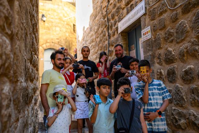 A group of children pose with their educators as they take pictures in the streets with film cameras during a workshop held as part of Fotohane Darkroom project in Mardin, southeastern Turkey, on June 14, 2026. In a dimly lit room illuminated by a pair of red lamps, eight-year-old Zeynep waits to see the photographs she has taken, now trapped as shadows and silhouettes on a roll of film. Originally from the southeastern province of Mardin, Zeynep is among eight children taking part in a two-month analog photography workshop for local and migrant youth near Turkey's borders with Iraq and Syria. The project, called Fotohane Darkroom, started in Mardin in 2024, by Kilic and Syrian photographer and educator Serbest Salih. In Turkish, Arabic, Kurdish and Persian, Fotohane means "house of photo", a name chosen by the children. (Photo by Yasin AKGUL / AFP)