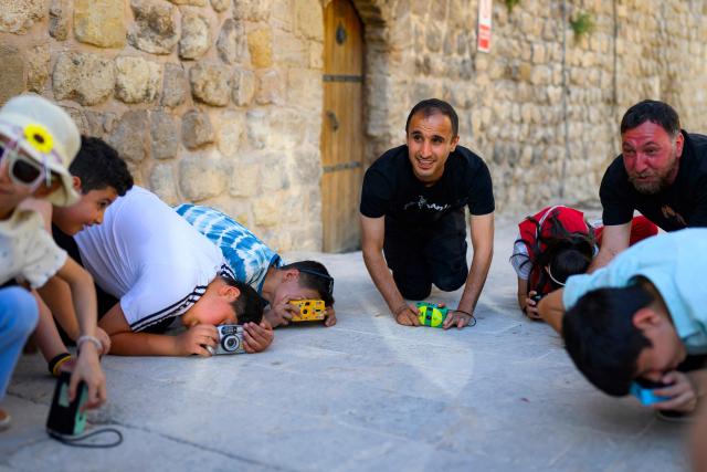 Syrian photographer, educator and co-founder of the project Serbest Salih (C) teaches children how to take pictures with film cameras in the streets during a workshop held as part of Fotohane Darkroom project in Mardin, southeastern Turkey, on June 14, 2026. In a dimly lit room illuminated by a pair of red lamps, eight-year-old Zeynep waits to see the photographs she has taken, now trapped as shadows and silhouettes on a roll of film. Originally from the southeastern province of Mardin, Zeynep is among eight children taking part in a two-month analog photography workshop for local and migrant youth near Turkey's borders with Iraq and Syria. The project, called Fotohane Darkroom, started in Mardin in 2024, by Kilic and Syrian photographer and educator Serbest Salih. In Turkish, Arabic, Kurdish and Persian, Fotohane means "house of photo", a name chosen by the children. (Photo by Yasin AKGUL / AFP)