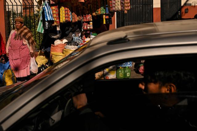 A car passes by as an Indigenous woman sells agricultural products on a street in downtown La Paz, Bolivia, on June 22, 2026. Bolivian police driving bulldozers cleared highways on June 21, 2026 and fuel trucks rolled into La Paz after the president declared a state of emergency to remove anti-government roadblocks that had paralyzed the Andean nation. (Photo by MARTIN BERNETTI / AFP)