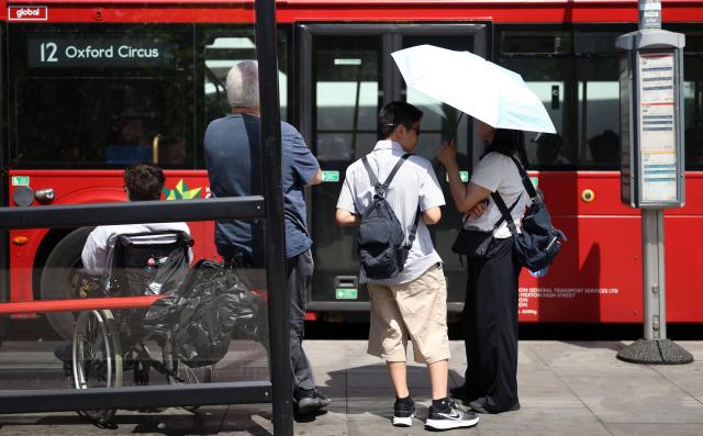 People use an umbrella to shelter from the direct sun, as they wait to board a red London bus, in London on June 23, 2026. (Photo by Toby Shepheard / AFP)