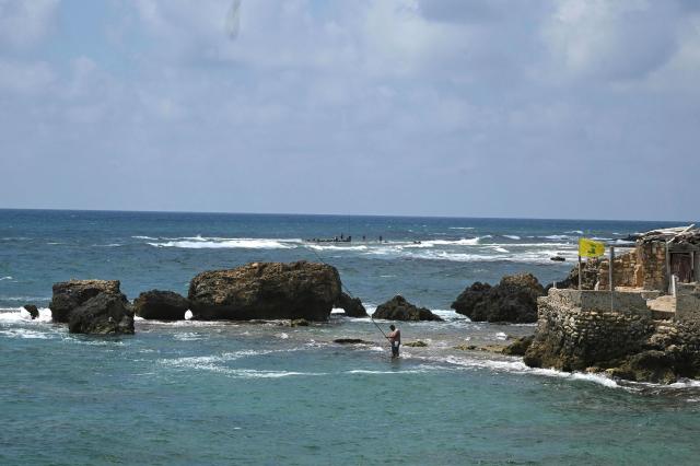 A yellow flag belonging to the Iran-backed Shia Muslim party Hezbollah flutters as a man fishes standing on the rocky shore of the Mediterranean Sea in the southern Lebanese coastal city of Tyre on June 23, 2026, days after the city saw numerous Israeli military strikes. On June 22, 2026, mediators Pakistan and Qatar said that Tehran and Washington had agreed to set up a "de-confliction cell" to limit flare-ups in Lebanon following talks in Switzerland on ending the wider Middle East war, which Tehran has linked to halting the parallel conflict in Lebanon. (Photo by Joseph EID / AFP) / 