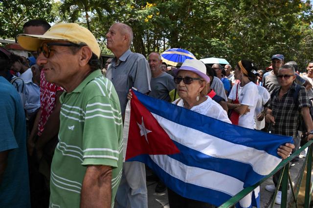 A woman holds a Cuban flag as she lines up in front of the Ministry of the Revolutionary Armed Forces to pay tribute to Commander Ramiro Valdés during his funeral in Havana on June 23, 2026. Ramiro Valdes Menendez, a close associate of brothers Fidel and Raul Castro and founder of the Cuban intelligence services, has died at the age of 94, Cuban President Miguel Diaz-Canel announced on June 21, 2026. (Photo by YAMIL LAGE / AFP)