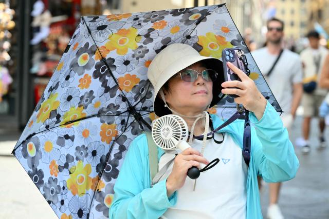 A tourist wearing a hat and holding a fan shields herself from the sun under an umbrella as she walks in Florence during a heatwave on June 24, 2026. (Photo by Alberto PIZZOLI / AFP)