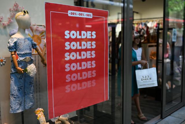 A customer walks past the entrance of a department store bearing a placard promoting French summer sales on the opening day in Beziers, Southern France, on June 24, 2026. The summer sales kick off on June 24, 2026, amid scorching heat, "a godsend" for air-conditioned department stores where consumers take refuge, but a source of concern for downtown shops, which fear purchases will shift to ultra-fast-fashion online platforms. (Photo by Gabriel BOUYS / AFP)