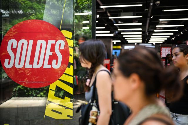 Pedestrians walk past the entrance of a department store bearing a placard promoting French summer sales on the opening day in Beziers, Southern France, on June 24, 2026. The summer sales kick off on June 24, 2026, amid scorching heat, "a godsend" for air-conditioned department stores where consumers take refuge, but a source of concern for downtown shops, which fear purchases will shift to ultra-fast-fashion online platforms. (Photo by Gabriel BOUYS / AFP)