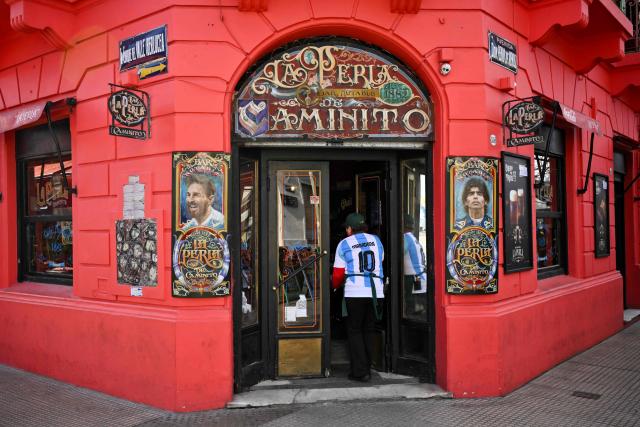 A woman wearing an Argentina national team jersey enters a restaurant decorated with images depicting Argentine football star Lionel Messi (L) and late Argentine football legend Diego Maradona in La Boca neighborhood, Buenos Aires on June 22, 2026, during the FIFA World Cup 2026. (Photo by Luis ROBAYO / AFP)