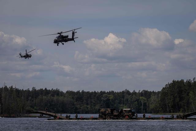 Two Polish Air Forces US-made AH-6h Apache helicopters fly over a Polish Army PFM Motorised Floating Bridge during a lake crossing, as troops from Poland and Lithuania take part in the "Bursztynowy Obronca-26" (Amber Defender) military exercise on the Orzysz military training ground, north-eastern Poland, on June 24, 2026. (Photo by Wojtek RADWANSKI / AFP)