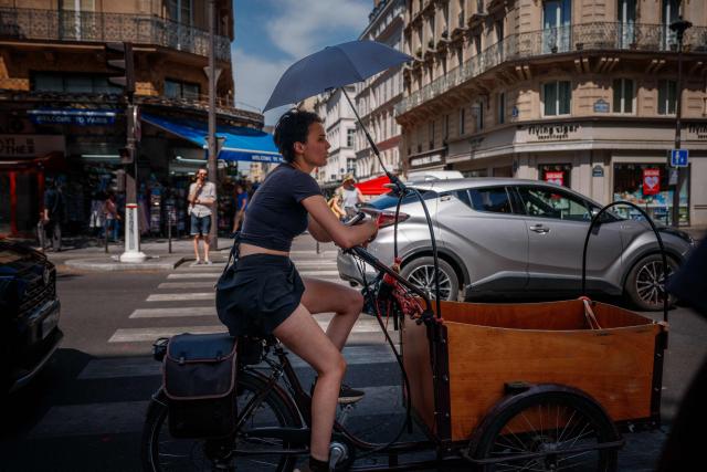 A woman protects herself from the sun under an umbrella as she rides a bicycle in Paris on June 25, 2026, as France experiences a heatwave. At least 101 million Europeans were expected to experience temperatures of over 35C on June 25, 2026, in a punishing and increasingly deadly heatwave hotter than weather in parts of Africa. France and Spain, among the countries worst hit, began counting the toll from the extreme temperatures, with children among the victims. (Photo by Dimitar DILKOFF / AFP)