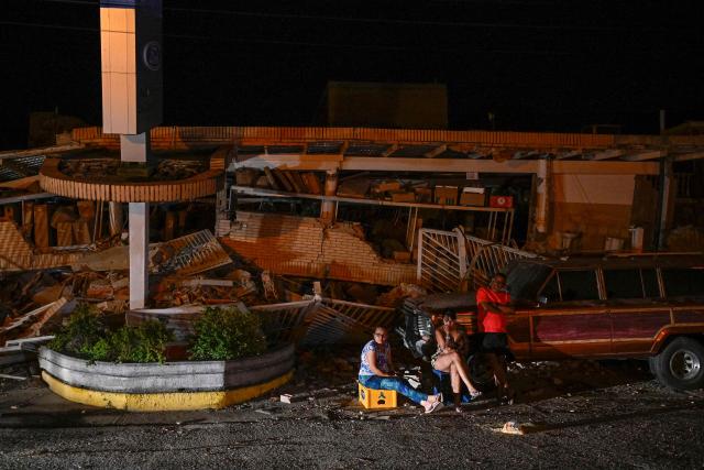 People sit in front of a damaged building following an earthquake in Catia La Mar, La Guaira state, some 30km north-west of Caracas, on June 25, 2026. A twin earthquake that was Venezuela's largest in over a century has killed at least 164 people and destroyed multiple buildings near the capital, where residents searched on June 25 for missing relatives. (Photo by Federico PARRA / AFP)