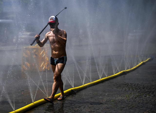 A man walks with a selfie stick past water hoses spilling water to provide relief from the summer heat as part of Colognes climate adaptation measures in Cologne, western Germany on June 25, 2026 during a heatwave in Europe. A heatwave sweeping Europe was starting to peak in Germany on June 25, 2026, with several open-air events cancelled and temperatures expected to top 40C through the weekend. Large parts of the country are already under "severe to extreme heat stress" with temperatures soaring to around 37C, the German Weather Service (DWD) said. (Photo by Ina FASSBENDER / AFP)