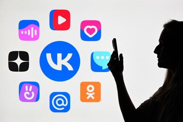 This illustration photograph shows a woman using a smartphone in front of the logos of Russian social media platform VK and Mail.ru group services displayed on a TV screen in Moscow on June 25, 2026. Apple has removed VKontakte, one of Russia's largest social networks, from its App Store, the Russian company said on June 25, 2026, with the Kremlin calling for an explanation from the California-based tech giant. VKontakte (VK) was launched in 2006 as Russia's answer to Facebook and has since grown to become one of the most popular websites and social media platforms in the country. (Photo by Igor IVANKO / AFP)