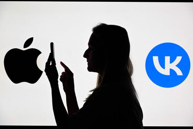 This illustration photograph shows a woman using a smartphone in front of the logos of Russian social media platform VK and Apple Inc displayed on a TV screen in Moscow on June 25, 2026. Apple has removed VKontakte, one of Russia's largest social networks, from its App Store, the Russian company said on June 25, 2026, with the Kremlin calling for an explanation from the California-based tech giant. VKontakte (VK) was launched in 2006 as Russia's answer to Facebook and has since grown to become one of the most popular websites and social media platforms in the country. (Photo by Igor IVANKO / AFP)