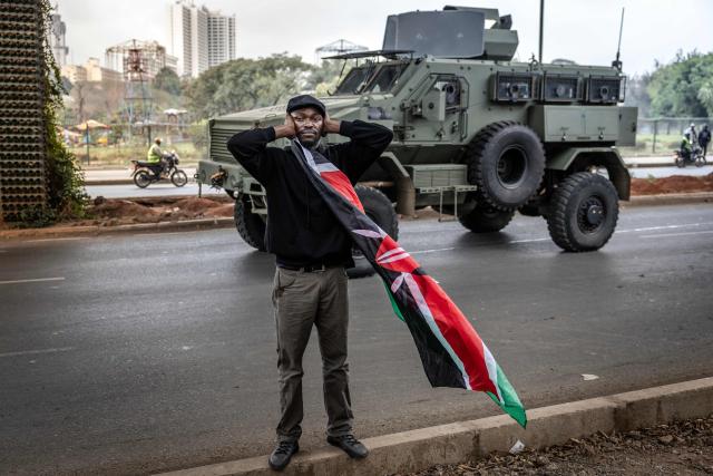 A protester covers her ears as a Kenyan police armored vehicle equipped with a sound cannon drives past while dispersing protesters during demonstrations marking the second anniversary of the deadly June 2024 youth-led anti-Finance Bill protests, which saw demonstrators storm Parliament, in Nairobi on June 25, 2026. Kenyan police put on a massive show of force on June 25 in a bid to prevent protests marking two years since a landmark Gen Z uprising. Thousands of Kenyans took to the streets in June 2024 against economic hardship and corruption, eventually storming parliament to force the cancellation of new taxes. (Photo by Luis TATO / AFP)