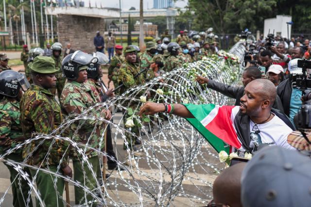 Kenyan police officers watch as activists place flowers on barbed-wire barrier set up to prevent them from gaining access to parliament precincts to lay floweres during demonstrations to mark the second anniversary of the deadly June 2024 youth-led anti-Finance Bill protests, which saw demonstrators storm Parliament, in Nairobi on June 25, 2026. Kenyan police put on a massive show of force on June 25 in a bid to prevent protests marking two years since a landmark Gen Z uprising. Thousands of Kenyans took to the streets in June 2024 against economic hardship and corruption, eventually storming parliament to force the cancellation of new taxes. (Photo by Tony KARUMBA / AFP)