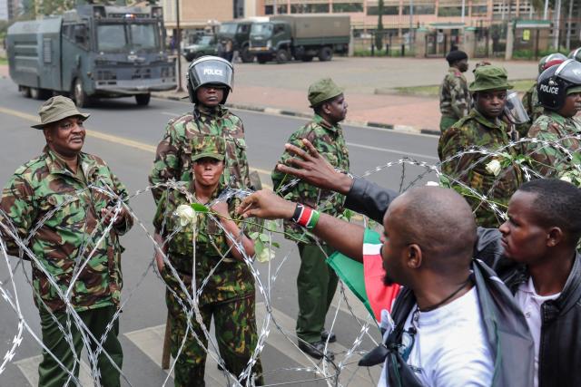 Kenyan police officers watch as activists place flowers on barbed-wire barrier set up to prevent them from gaining access to parliament precincts to lay floweres during demonstrations to mark the second anniversary of the deadly June 2024 youth-led anti-Finance Bill protests, which saw demonstrators storm Parliament, in Nairobi on June 25, 2026. Kenyan police put on a massive show of force on June 25 in a bid to prevent protests marking two years since a landmark Gen Z uprising. Thousands of Kenyans took to the streets in June 2024 against economic hardship and corruption, eventually storming parliament to force the cancellation of new taxes. (Photo by Tony KARUMBA / AFP)