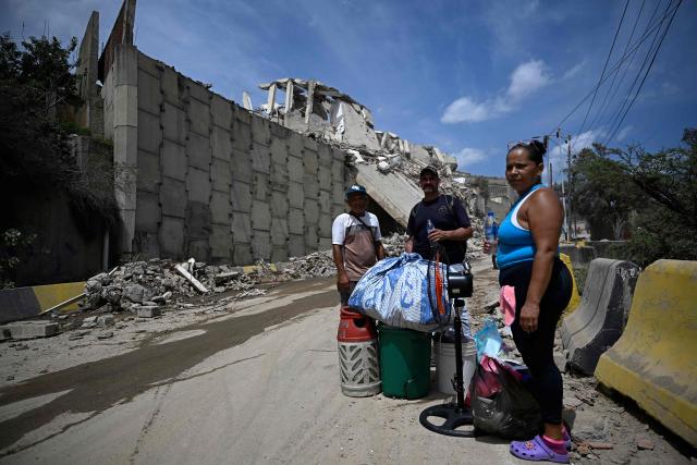 Residents stand beside salvaged belongings on a street near a collapsed structure in Catia La Mar, La Guaira state, about 30 km northwest of Caracas, on June 25, 2026, following an earthquake. A twin earthquake that was Venezuela's largest in over a century has killed at least 164 people and destroyed multiple buildings near the capital, where residents searched on June 25 for missing relatives. (Photo by Federico PARRA / AFP)