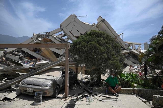 A man sits near a damaged car outside a collapsed structure following an earthquake in Los Corales, La Guaira state, about 38 km northwest of Caracas, on June 25, 2026. A twin earthquake that was Venezuela's largest in over a century has killed at least 188 people and destroyed multiple buildings near the capital, where residents searched on June 25 for missing relatives. (Photo by Federico PARRA / AFP)