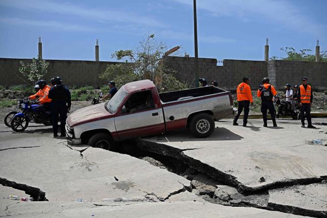 Locals ride past police officers standing next to a vehicle stranded in a cracked street following a twin earthquake in Los Corales, La Guaira state, about 38 km northwest of Caracas, on June 25, 2026. The death toll from twin quakes, measured at magnitudes 7.2 and 7.5, has risen to at least 188, with more than 1,520 others injured, top lawmaker Jorge Rodriguez reported, while residents continue searching for missing relatives. (Photo by Federico PARRA / AFP)