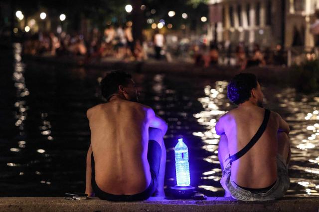 Two youths sit along the bank of the Canal Saint-Martin to cool down at nightfall, next to an illuminated water bottle, as France experiences a heatwave, in Paris on June 25, 2026. Due to the ongoing heatwave sweeping across France - the Bassin des Recollets - a portion of the Canal Saint-Martin in the French capital has been opened for swimming from June 17. Authorities said hospitals in Paris and its surrounding region were "saturated" and decided to ban alcohol consumption in the streets and public spaces from June 26 at noon, as well as alcohol sales from 6:00 pm. (Photo by Ludovic MARIN / AFP)