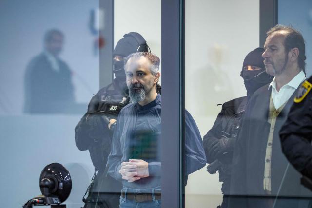Saudi Arabian defendant Taleb Jawad al-Abdulmohsen (C) stands next to a judicial security officer as presiding judge Dirk Sternberg (L) is reflected in a bulletproof glass box in the temporary courtroom in Magdeburg, eastern Germany, before al-Abdulmohsen's trial for driving an SUV through a crowd at a Christmas market. The court sentenced to life in jail the Saudi psychiatrist who drove an SUV through a crowded Christmas market in 2024, killing six people and injuring more than 300. (Photo by Ronny HARTMANN / AFP)