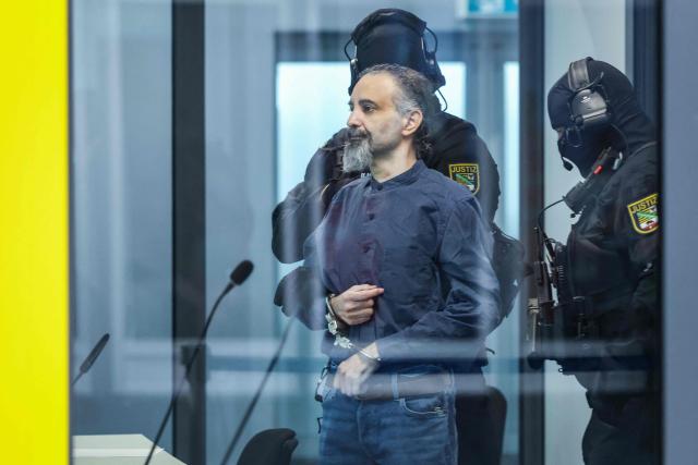 Saudi Arabian defendant Taleb Jawad al-Abdulmohsen (L) stands next to a judicial security officer as he waits inside a bulletproof glass box in the temporary courtroom in Magdeburg, eastern Germany, before his trial for driving an SUV through a crowd at a Christmas market. The court sentenced to life in jail the Saudi psychiatrist who drove an SUV through a crowded Christmas market in 2024, killing six people and injuring more than 300. (Photo by Ronny HARTMANN / AFP)