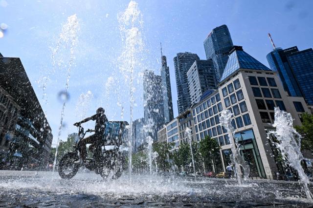 A man rides his bicycle through a fountain outside the banking district in central Frankfurt am Main, western Germany, on June 26, 2026 with air temperature reaching 39 degrees Celsius. A heatwave sweeping Europe was starting to peak in Germany, with several open-air events cancelled and temperatures expected to top 40C through the weekend. Large parts of the country are already under "severe to extreme heat stress", the German Weather Service (DWD) said. (Photo by Kirill KUDRYAVTSEV / AFP)