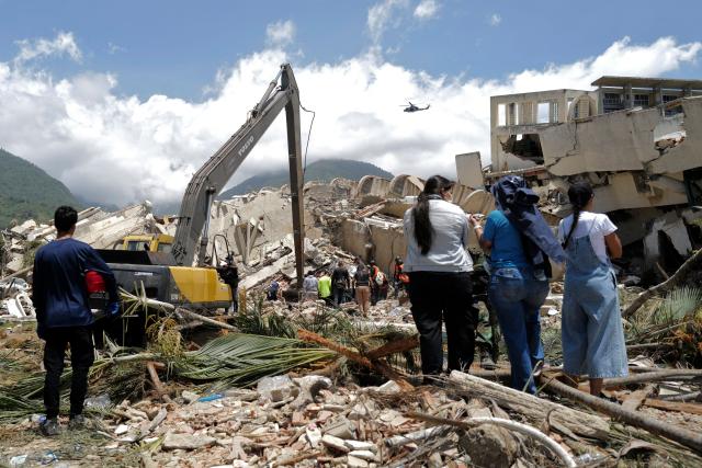 Locals stand as a backhoe loader removes rubble searching for survivors of the twin earthquakes, in Caraballeda, La Guaira State, some 40 km northeast of Caracas, on June 27, 2026. Three days after powerful tremors of magnitude 7.2 and 7.5 killed 1,430 people and left more than 50,000 missing, fury with perceived governmental inaction is mounting -- as is the feeling of helplessness. (Photo by Mauricio VALENZUELA / AFP)