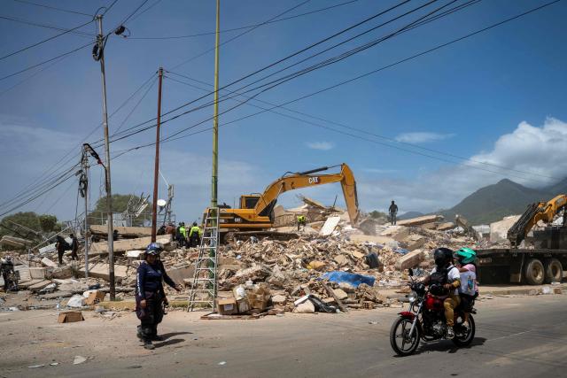 Locals ride past heavy machinery removing debris of a collapsed building in Caraballeda, La Guaira state, Venezuela, on June 27, 2026, following twin earthquakes. Nearly 1,000 people have been killed and more than 50,000 remain missing after two consecutive earthquakes in Venezuela, where growing anger has been reported over the lack of official aid to swiftly rescue survivors. (Photo by Miguel MEDINA / POOL / AFP)