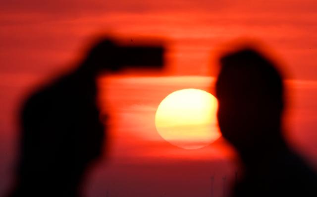 A person takes a photo as the sun going up on early June 28, 2026, over the Drachenberg mountain in Berlin, as the German capital is expected to reach teperatures around 40 degrees Celsius during a heatwave in Europe. Tens of millions are braving a weekend of extreme temperatures in Europe as a deadly heatwave moves eastwards, with some countries announcing rising death tolls and health services warning of saturation. While some mild relief is expected Sunday in western Europe, German forecasters are warning that more temperature records could still be broken over the weekend as eastern countries issue a slew of red alerts for the coming days. (Photo by RALF HIRSCHBERGER / AFP)