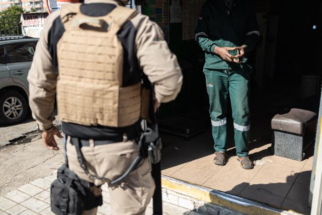 A private security officer questions a man as he holds his passport during a joint patrol in the Johannesburg CBD on June 28, 2026. The operation, conducted alongside members of the South African Police Service (SAPS) (unseen) forms part of Operation Shanela, a policing initiative targeting the inner city. The coordinated effort sees SAPS working in closer operational alignment with private security firms, such as Professional Protection Security (PPS). Focus areas include crime prevention, immigration compliance checks, and on-the-ground intelligence gathering. (Photo by EMMANUEL CROSET / AFP)