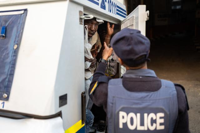 TOPSHOT - A South African Police Service (SAPS) officer gestures to detained men inside a police van during a joint patrol in the Johannesburg CBD on June 28, 2026. The operation, conducted alongside members of the South African Police Service (SAPS) (unseen) forms part of Operation Shanela, a policing initiative targeting the inner city. The coordinated effort sees SAPS working in closer operational alignment with private security firms, such as Professional Protection Security (PPS). Focus areas include crime prevention, immigration compliance checks, and on-the-ground intelligence gathering. (Photo by EMMANUEL CROSET / AFP)