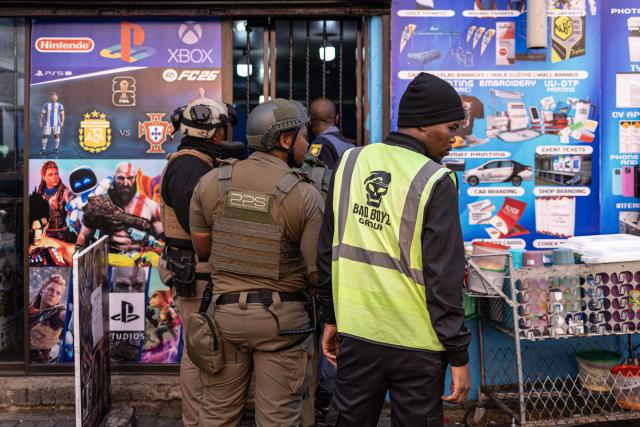 Private security officers along with a South African Police Service (SAPS) officer prepare to enter a shop during a joint patrol in the Johannesburg CBD on June 28, 2026. The operation, conducted alongside members of the South African Police Service (SAPS) (unseen) forms part of Operation Shanela, a policing initiative targeting the inner city. The coordinated effort sees SAPS working in closer operational alignment with private security firms, such as Professional Protection Security (PPS). Focus areas include crime prevention, immigration compliance checks, and on-the-ground intelligence gathering. (Photo by EMMANUEL CROSET / AFP)