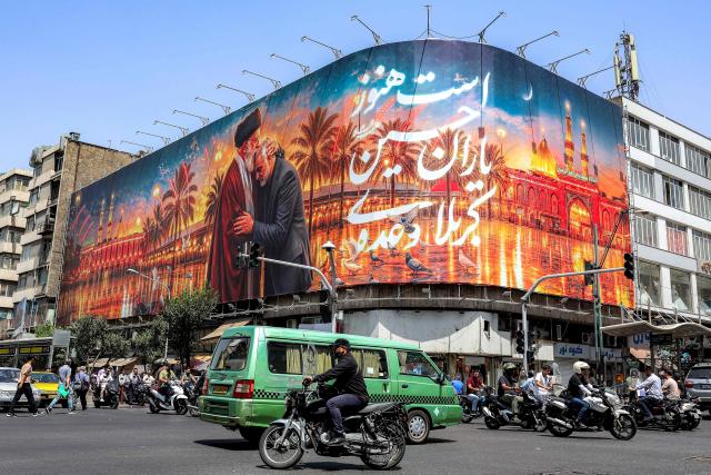 Motorists drive vehicles past a billboard depicting Iran's slain supreme leader Ali Khamenei kissing the slain Islamic revolutionary guards commander Qasem Soleimani near the shrine of Imam Hussein Iraq's holy city of Karbala, displayed on the facade of a building in Tehran on June 29, 2026. Iran had on June 23 announced three days of public holidays, from July 4 to 6, in the capital Tehran for late supreme leader Ayatollah Ali Khamenei's funeral ceremonies, state television reported. He will finally be laid to rest on July 9 in the northeastern holy city of Mashhad, his birthplace. Khamenei was killed in a strike on the first day of the US-Israeli war against the Iran on February 28. (Photo by AFP) / 