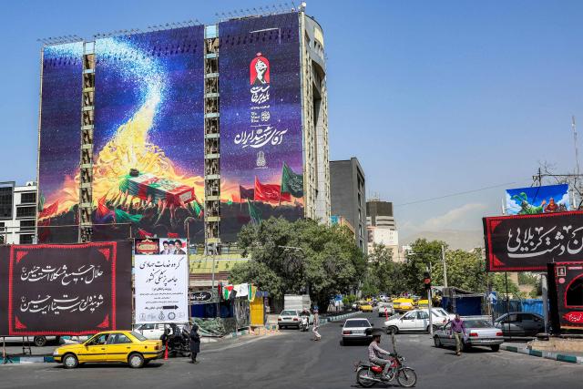 Motorists drive vehicles past a mural displayed on the facade of a building depicts late figures aligned with the Iranian government, such as late President Ebrahim Raisi, slain Islamic revolutionary guards commander Qasem Soleimani, slain Lebanese Hezbollah leader Hassan Nasrallah, and others, all carrying the flag-draped casket of Iran's slain supreme leader Ali Khamenei who was killed in a strike on the first day of the US-Israeli war against the Iran, at Vanak Square in Tehran on June 29, 2026. Iran had on June 23 announced three days of public holidays, from July 4 to 6, in the capital Tehran for late supreme leader Ayatollah Ali Khamenei's funeral ceremonies, state television reported. He will finally be laid to rest on July 9 in the northeastern holy city of Mashhad, his birthplace. (Photo by AFP) / 