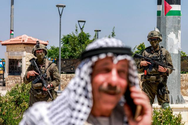 A Palestinian man speaks on a phone near Israeli army soldiers standing guard, during an official visit by a group of foreign diplomats accredited by the Palestinian Authority at the archaeological site of Sebastia, north of Nablus in the occupied West Bank, on June 29, 2026. (Photo by Jaafar ASHTIYEH / AFP)