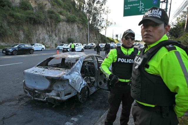 Police remain next to a burned car, which had allegedly transported the people responsible for planting a bomb, in the outskirts of Quito on June 29, 2026. Ecuadoran police conducted a controlled explosion of a bomb planted outside the Ministry of Agriculture shattering windows there. It was the second time in five days that a bomb has been found in the capital Quito, and comes as Ecuador grapples with rising violence by drug cartels. (Photo by Rodrigo BUENDIA / AFP)