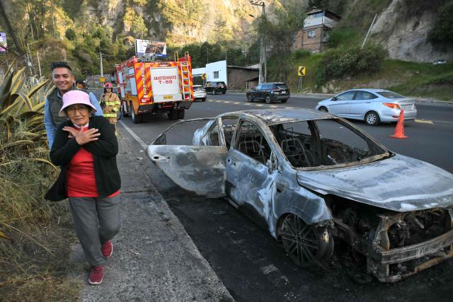 People walk past a burned car, which had allegedly transported the people responsible for planting a bomb, in the outskirts of Quito on June 29, 2026. Ecuadoran police conducted a controlled explosion of a bomb planted outside the Ministry of Agriculture shattering windows there. It was the second time in five days that a bomb has been found in the capital Quito, and comes as Ecuador grapples with rising violence by drug cartels. (Photo by Rodrigo BUENDIA / AFP)