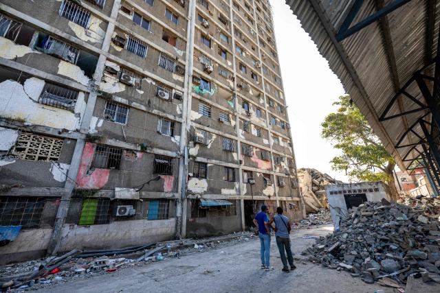 Locals stand next to a dammaged building in Catia La Mar, La Guaira state, Venezuela, on June 29, 2026. Hopes were fading on June 29, 2026 of finding survivors more than four days after powerful twin earthquakes struck Venezuela, as residents grow increasingly frustrated with the government's response to the disaster that has killed at least 1,450 people and left tens of thousands unaccounted for. (Photo by Miguel MEDINA / POOL / AFP)