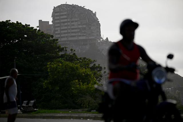 A motorcyclist rides past as a heavily damaged multi-storey building stands in the background in the Playa Verde sector of Catia La Mar, La Guaira state, Venezuela, on June 29, 2026, following twin earthquakes. Hopes were fading of finding survivors more than four days after powerful twin earthquakes struck Venezuela, as residents grow increasingly frustrated with the government's response to the disaster that has killed -up to now- more than 1,700 people and left tens of thousands unaccounted for. (Photo by Federico PARRA / POOL / AFP)