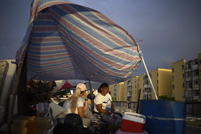 Displaced residents take shelter under a makeshift tarp set up over a vehicle near apartment buildings in the Playa Verde sector of Catia La Mar, La Guaira state, Venezuela, on June 29, 2026, following twin earthquakes. Hopes were fading of finding survivors more than four days after powerful twin earthquakes struck Venezuela, as residents grow increasingly frustrated with the government's response to the disaster that has killed -up to now- more than 1,700 people and left tens of thousands unaccounted for. (Photo by Federico PARRA / POOL / AFP)