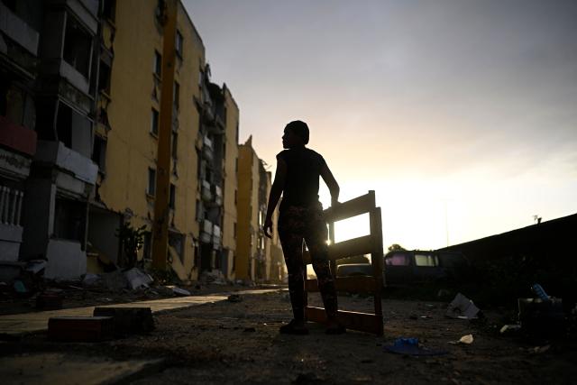 A woman stands holding a wooden structure amid debris along a damaged street near apartment buildings in the Playa Verde sector of Catia La Mar, La Guaira state, Venezuela, on June 29, 2026, following twin earthquakes. Hopes were fading of finding survivors more than four days after powerful twin earthquakes struck Venezuela, as residents grow increasingly frustrated with the government's response to the disaster that has killed -up to now- more than 1,700 people and left tens of thousands unaccounted for. (Photo by Federico PARRA / POOL / AFP)